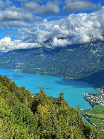 A picturesque landscape featuring a vibrant turquoise lake surrounded by lush green forests. Majestic mountains rise in the background, partially covered by thick, fluffy clouds. A small village is visible along the shoreline, adding charm to the serene natural setting.