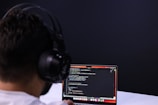 A person with headphones is engaged in coding on a laptop. The screen displays lines of code, possibly written in a programming language like JavaScript or Python. The setting has a minimalistic and focused atmosphere, with a dark background.
