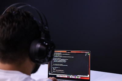 A person with headphones is engaged in coding on a laptop. The screen displays lines of code, possibly written in a programming language like JavaScript or Python. The setting has a minimalistic and focused atmosphere, with a dark background.