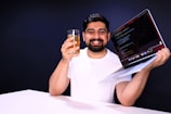 A person with short dark hair and a beard is holding a glass with a yellow beverage in one hand and a laptop displaying code in the other. The individual is smiling and wearing a white shirt. The background is a plain dark color.