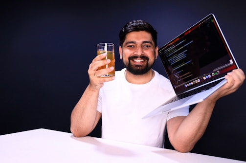 A person with short dark hair and a beard is holding a glass with a yellow beverage in one hand and a laptop displaying code in the other. The individual is smiling and wearing a white shirt. The background is a plain dark color.