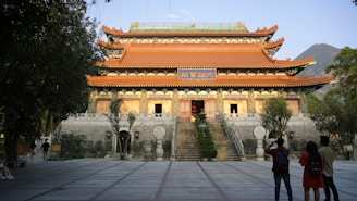 A traditional Chinese temple with ornate architectural features is surrounded by trees. The temple has a tiled roof with decorative eaves and a prominent sign with Chinese characters above the entrance. Several people are standing in the foreground, admiring the structure. The scene is set in an open courtyard with steps leading up to the temple.