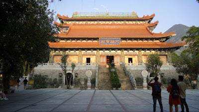 A traditional Chinese temple with ornate architectural features is surrounded by trees. The temple has a tiled roof with decorative eaves and a prominent sign with Chinese characters above the entrance. Several people are standing in the foreground, admiring the structure. The scene is set in an open courtyard with steps leading up to the temple.