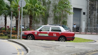 A red taxi is parked on a street corner near a small patch of grass and a group of palm trees. The surroundings include a modern building with a gray facade and a partially visible person in the background wearing a hat.