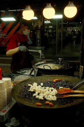 A street food vendor wearing a mask and gloves prepares food in a bustling indoor market. The scene is lit by warm overhead lights, and there's a large round pan filled with rice cakes and some red sauce. Stacks of paper cups are visible on the side, and background shows a reflective glass window with people walking past and various shop signs.