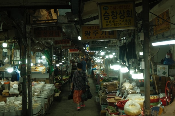 A bustling indoor market scene with narrow aisles lined with various stalls. Each stall is filled with a diverse range of goods, including bags of grains, spices, and fresh produce. Bright overhead lights illuminate the market, creating a lively and busy atmosphere. Several people, including a woman carrying a bag, walk through the aisle, browsing the offerings.