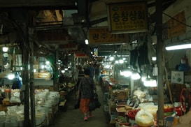 A bustling indoor market scene with narrow aisles lined with various stalls. Each stall is filled with a diverse range of goods, including bags of grains, spices, and fresh produce. Bright overhead lights illuminate the market, creating a lively and busy atmosphere. Several people, including a woman carrying a bag, walk through the aisle, browsing the offerings.