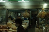 A dimly lit indoor market stall displays various dried goods in plastic bags. The interior is decorated with hanging bags of dried seafood, and shelves are stocked with numerous packages of snacks and grains. A person wearing a mask is visible in the foreground, partially obscured.
