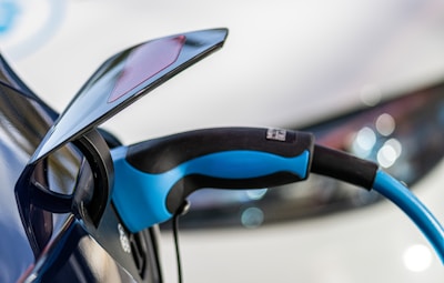 A close-up view of an electric vehicle charging port with a blue and black charging cable plugged into it. The background features a blurred image of a car, highlighting the charging connection.