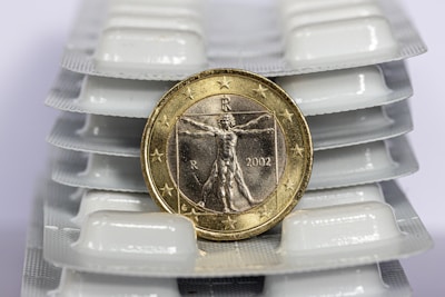 A coin with the Vitruvian Man design is placed upright against a backdrop of white pill blister packs. The coin is detailed with stars along its edges and is dated 2002.