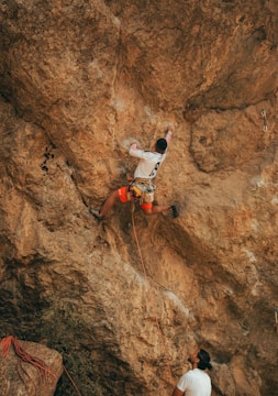 A person wearing a white long-sleeve shirt and orange shorts is climbing a steep, rocky cliff. Another individual on the ground is holding a safety rope, assisting in belaying the climber. The rock surface is rugged and textured, with various shades of brown.