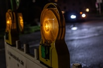 A nighttime scene featuring a road barrier with illuminated warning lights. The lights emit a warm glow, contrasting with the dark surroundings. In the background, a car with headlights on approaches, casting reflections on the wet pavement.