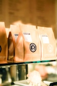 Several brown paper coffee bags are lined up on a glass shelf, each with a label indicating the brand 'Coffee & Water Lab' and specific details about the coffee. The labels feature a graphic of a coffee dripper. The scene is warmly lit, giving a cozy and inviting ambiance.