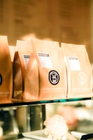 Several brown paper coffee bags are lined up on a glass shelf, each with a label indicating the brand 'Coffee & Water Lab' and specific details about the coffee. The labels feature a graphic of a coffee dripper. The scene is warmly lit, giving a cozy and inviting ambiance.