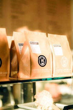 Several brown paper coffee bags are lined up on a glass shelf, each with a label indicating the brand 'Coffee & Water Lab' and specific details about the coffee. The labels feature a graphic of a coffee dripper. The scene is warmly lit, giving a cozy and inviting ambiance.