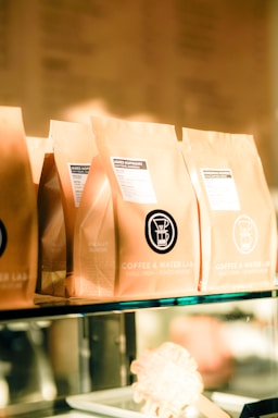 Several brown paper coffee bags are lined up on a glass shelf, each with a label indicating the brand 'Coffee & Water Lab' and specific details about the coffee. The labels feature a graphic of a coffee dripper. The scene is warmly lit, giving a cozy and inviting ambiance.