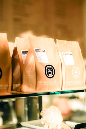 Several brown paper coffee bags are lined up on a glass shelf, each with a label indicating the brand 'Coffee & Water Lab' and specific details about the coffee. The labels feature a graphic of a coffee dripper. The scene is warmly lit, giving a cozy and inviting ambiance.