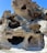 A complex formation of weathered rock with multiple openings and cavities, featuring smooth surfaces and a rugged texture. The stone appears to be light beige with darker shadows inside the openings, set against a clear blue sky.