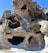 A complex formation of weathered rock with multiple openings and cavities, featuring smooth surfaces and a rugged texture. The stone appears to be light beige with darker shadows inside the openings, set against a clear blue sky.