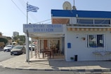 A Greek taverna with a sign in Greek, featuring an outdoor seating area with wooden tables and chairs. The building is white with blue accents and has a Greek flag fluttering on a pole nearby. Cars are parked and driving on the adjacent street, and there is a mix of residential and commercial buildings in the background.