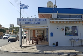 A Greek taverna with a sign in Greek, featuring an outdoor seating area with wooden tables and chairs. The building is white with blue accents and has a Greek flag fluttering on a pole nearby. Cars are parked and driving on the adjacent street, and there is a mix of residential and commercial buildings in the background.