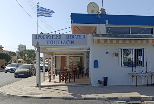 A Greek taverna with a sign in Greek, featuring an outdoor seating area with wooden tables and chairs. The building is white with blue accents and has a Greek flag fluttering on a pole nearby. Cars are parked and driving on the adjacent street, and there is a mix of residential and commercial buildings in the background.