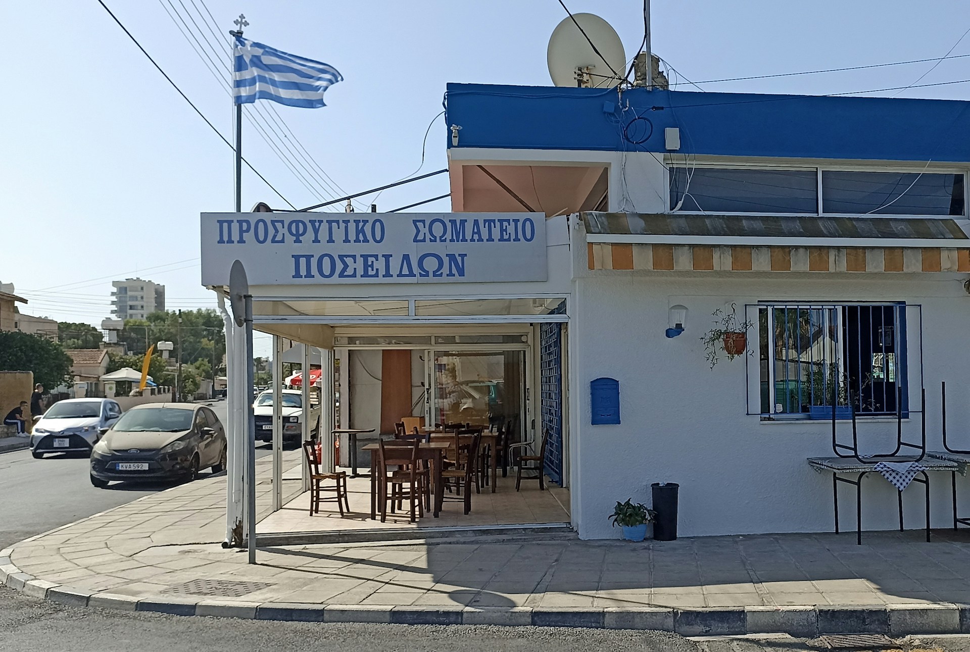 A Greek taverna with a sign in Greek, featuring an outdoor seating area with wooden tables and chairs. The building is white with blue accents and has a Greek flag fluttering on a pole nearby. Cars are parked and driving on the adjacent street, and there is a mix of residential and commercial buildings in the background.
