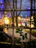 A vibrant holiday scene with brightly lit decorations including Christmas lights and illuminated trees. In the foreground, a wooden bench is placed on a cobblestone path surrounded by greenery. The background reveals a festive atmosphere with colorful lights reflecting off a body of water, and a ferris wheel or amusement ride visible in the distance.