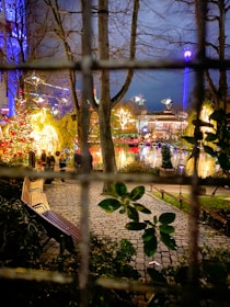A vibrant holiday scene with brightly lit decorations including Christmas lights and illuminated trees. In the foreground, a wooden bench is placed on a cobblestone path surrounded by greenery. The background reveals a festive atmosphere with colorful lights reflecting off a body of water, and a ferris wheel or amusement ride visible in the distance.