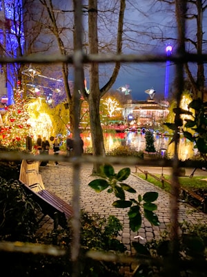 A vibrant holiday scene with brightly lit decorations including Christmas lights and illuminated trees. In the foreground, a wooden bench is placed on a cobblestone path surrounded by greenery. The background reveals a festive atmosphere with colorful lights reflecting off a body of water, and a ferris wheel or amusement ride visible in the distance.