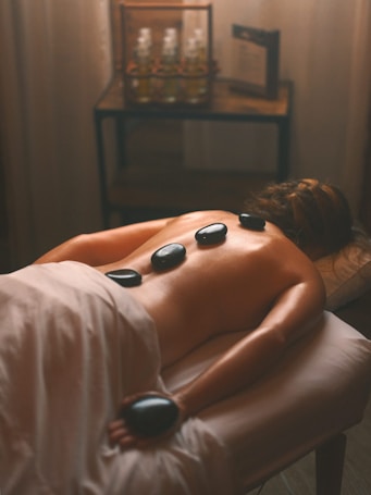 A serene spa setting featuring a person lying face down on a massage table covered with a white sheet. Several smooth black stones are placed along their back for a hot stone massage. Soft, ambient lighting creates a warm atmosphere, with bottles of oil visible on a wooden table in the background.