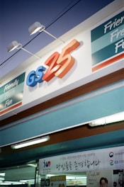 A convenience store sign with the logo GS25 prominently displayed in blue and orange letters. Below the logo, a banner with Korean text is visible. The storefront is brightly lit with fluorescent lights, and phrases like 'Friendly Fresh Fun' are featured on the facade.