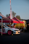 A white sports car with pink rims, marked with the number 81 and logos on the side, is parked under a canopy. The area has multiple tents and a yellow utility vehicle in the background. There are people standing nearby, and the scene is set in an outdoor location during daytime.