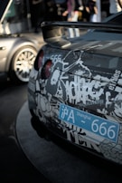 A close-up view of the rear of a car adorned with a complex pattern of graffiti-style graphics. The back of the car displays a blue Shanghai license plate with the number 666. The taillights are red and round, contrasting with the monochrome decals covering the vehicle. In the blurred background, parts of another car and out-of-focus people can be seen, suggesting an urban setting or automotive event.