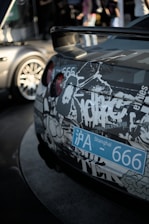 A close-up view of the rear of a car adorned with a complex pattern of graffiti-style graphics. The back of the car displays a blue Shanghai license plate with the number 666. The taillights are red and round, contrasting with the monochrome decals covering the vehicle. In the blurred background, parts of another car and out-of-focus people can be seen, suggesting an urban setting or automotive event.