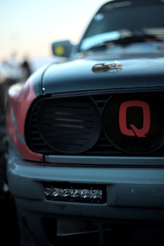 Close-up of the front part of a car featuring a prominent grille with the letter 'Q' in red. The grill is framed by sleek lines and geometric shapes. The focus is on the detail of the car's design, with the background blurred.
