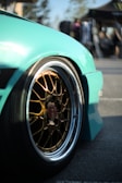 A close-up view of a car wheel with a glossy turquoise fender. The wheel features intricate gold-colored alloy rims with a branded center cap. The background is blurred, suggesting an outdoor setting with people and a tent.