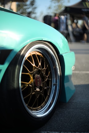 A close-up view of a car wheel with a glossy turquoise fender. The wheel features intricate gold-colored alloy rims with a branded center cap. The background is blurred, suggesting an outdoor setting with people and a tent.