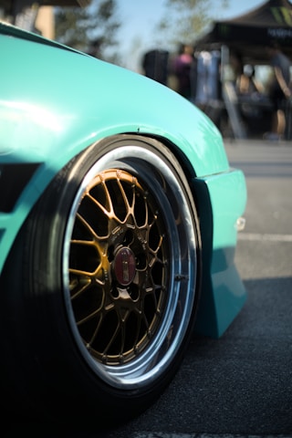 A close-up view of a car wheel with a glossy turquoise fender. The wheel features intricate gold-colored alloy rims with a branded center cap. The background is blurred, suggesting an outdoor setting with people and a tent.