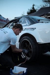 A person is kneeling next to a white car, appearing to be adjusting or inspecting the wheel. The car has visible branding and decals on its side. The setting appears to be outdoors, possibly at a racing or automotive event judging by the presence of tents and other vehicles in the background.