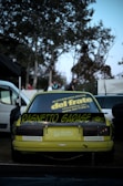 A yellow sports car is parked outdoors with a custom spoiler featuring the text 'Ragnetto Garage.' The rear windshield displays various decals, including one promoting a demolition company. The scene is set against a backdrop of trees, with a few people visible in the distance. Surrounding vehicles and objects, such as tires, can be seen.