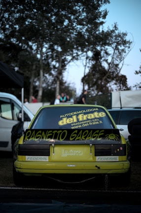 A yellow sports car is parked outdoors with a custom spoiler featuring the text 'Ragnetto Garage.' The rear windshield displays various decals, including one promoting a demolition company. The scene is set against a backdrop of trees, with a few people visible in the distance. Surrounding vehicles and objects, such as tires, can be seen.