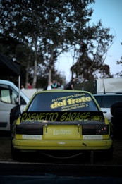 A yellow sports car is parked outdoors with a custom spoiler featuring the text 'Ragnetto Garage.' The rear windshield displays various decals, including one promoting a demolition company. The scene is set against a backdrop of trees, with a few people visible in the distance. Surrounding vehicles and objects, such as tires, can be seen.