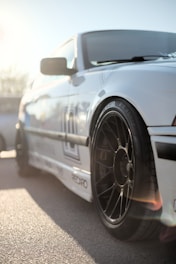 A close-up view of a white sports car with black rims, taken from a low angle. The sunlight is beaming, creating lens flare and highlighting the car's glossy exterior. There are other vehicles in the blurred background, suggesting an outdoor setting in daylight.