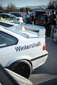Several cars are parked in an outdoor area, with a white car featuring the text 'Wintershall' prominently displayed on its rear side. People stand and walk around, engaging in conversation, set against a backdrop of trees and distant mountains.