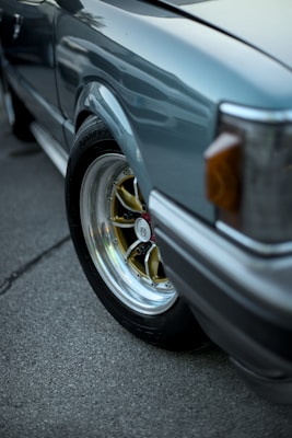 Close-up view of a car's wheel and part of the fender. The wheel features a polished silver rim with a gold center and black tire. The car body is painted in a metallic hue with visible reflections, and the headlight area has an amber detail. The ground consists of a textured, dark gray asphalt.
