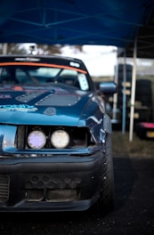 A close-up view of the front of a modified racing car with a damaged bumper. The vehicle has a blue exterior with visible sticker decals and customized headlights. The background includes a set of stacked tires and a blue canopy overhead, suggesting a motorsport event setting.