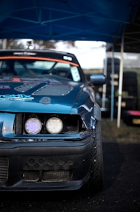A close-up view of the front of a modified racing car with a damaged bumper. The vehicle has a blue exterior with visible sticker decals and customized headlights. The background includes a set of stacked tires and a blue canopy overhead, suggesting a motorsport event setting.