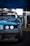 A close-up view of the front of a modified racing car with a damaged bumper. The vehicle has a blue exterior with visible sticker decals and customized headlights. The background includes a set of stacked tires and a blue canopy overhead, suggesting a motorsport event setting.