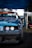 A close-up view of the front of a modified racing car with a damaged bumper. The vehicle has a blue exterior with visible sticker decals and customized headlights. The background includes a set of stacked tires and a blue canopy overhead, suggesting a motorsport event setting.